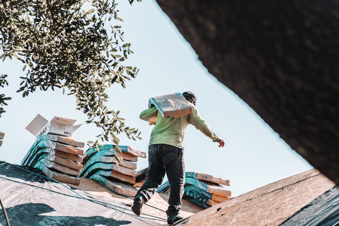 A professional roofer in safety gear meticulously installs new architectural shingles on a residential roof on a bright, sunny day.
