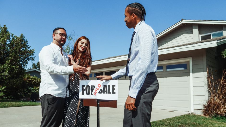 A real estate agent hands keys to a happy couple in front of a beautiful modern home, symbolizing a successful and fast sale.